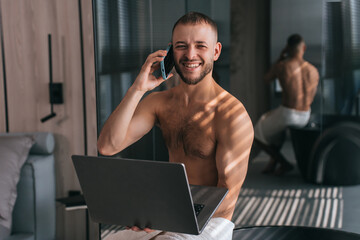 Young bearded caucasian guy sitting on bath with towel around hips talks by phone holds laptop smiles wide at bathroom. Successful young businessman at hotel room talks with partner. Wealthy life.