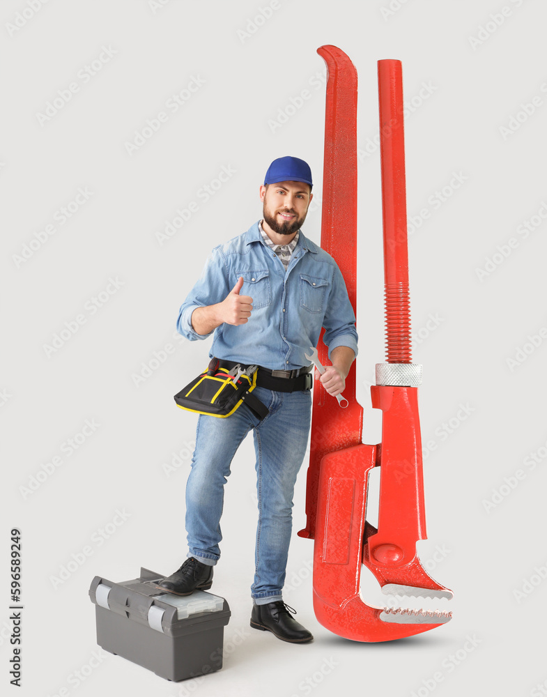 Poster handsome plumber with big pipe wrench showing thumb-up on light background