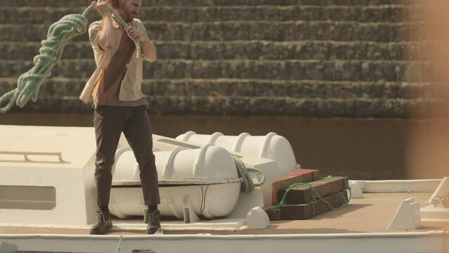 Full length shot of Caucasian sailor carrying rope walking along boat deck lit by sunlight