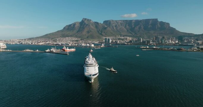 Aerial zoom in. Tugs escorting a Cruise ship out Cape Town harbour, Spectacular Table Mountain in the background.