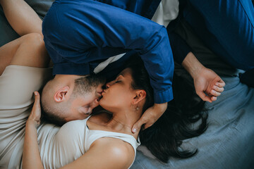 Top view on kissing young couple laying on bed at home. Lovers embracing each other at bedroom. Romance concept. Husband kissing wife  at home. Relationship, love.