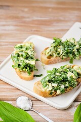 Open sandwiches with spring salad of wild garlic and boiled eggs on a white ceramic board on a wooden background. Using wild plants for food