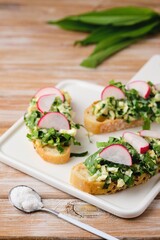 Open sandwiches with spring salad of wild garlic, boiled eggs and radish slices on a white ceramic board on a wooden background. Using wild plants for food