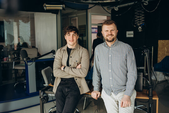 Two college podcasters laughing and having a good time in a studio. Two happy young men co-hosting a live audio broadcast. Two male content creators recording an internet podcast.