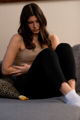 Depressed young woman sitting on the couch. Brunette girl having period pain and touching her belly