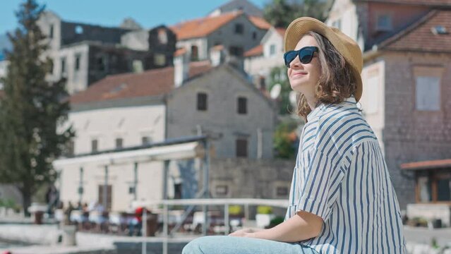 A young Caucasian woman in a hat uses her phone while sitting against the background of an old European seaside town
