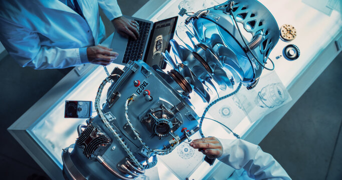 Top Down View of Two Engineers Discussing Work, Talking, Using Laptop Computer and Checking Blueprints in Jet Motor Manufacturing Facility. Scientists Researching Innovative Turbine Engine