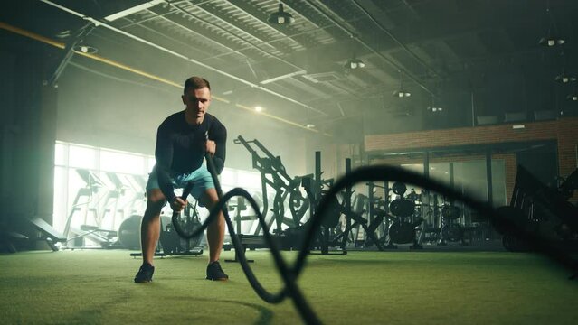 Young well-built man in black long sleeve compression shirt and blue shorts doing alternative waves on battle rope in gym. Cardio area in the background. High quality 4k footage