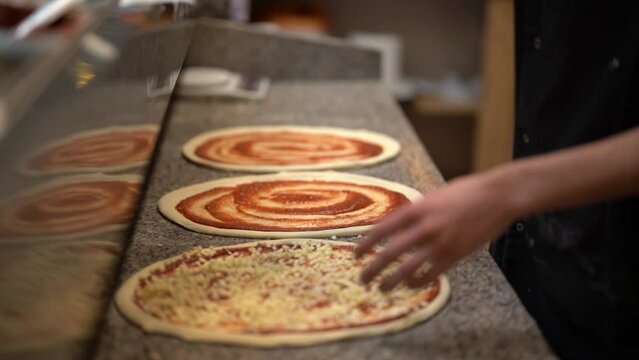 Side angle of unrecognizable male pizza chef decorating three pizzas with grated cheese standing in front of grey marble kitchen table in pizzeria 