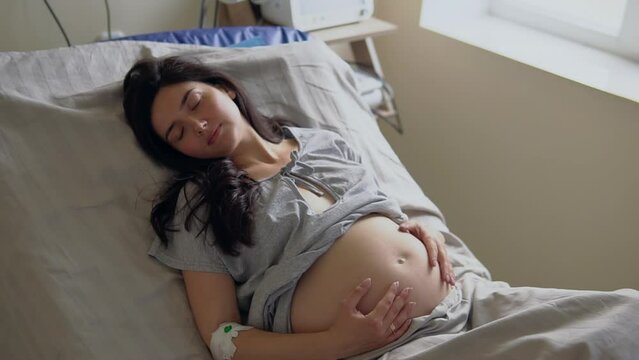 Brunette woman lies on bed stroking abdomen in ward