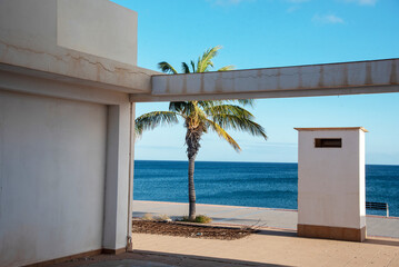 Arquitectura simple y blanca típica de la Fuerteventura turística, en el medio una ventana al mar turquesa y una palmera tropical movida por el viento en el pueblo de Tarajalejo en las islas Canarias