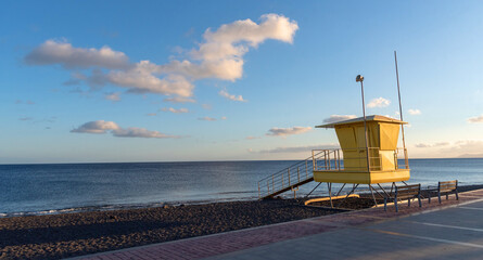 Vista panorámica de la playa de Tarajalejo de arena negra y aguas tranquilas y cristalinas al atardecer, con un puesto de socorrista amarillo en el lago y el paseo marítimo al lado en Fuerteventura