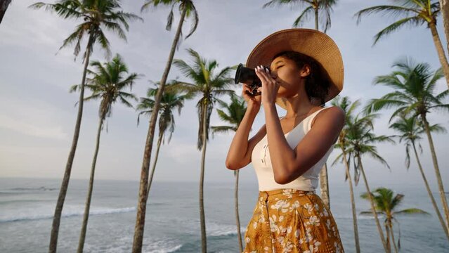 African american woman takes photos with camera on tropical island. Female multiracial photographer tourist explores ocean view location on vacation. Black girl taking pictures on sunrise.