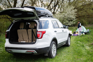 Big SUV car with open trunk and roof rack box against happy young family having fun and enjoying outdoor on picnic blanket at garden spring park.