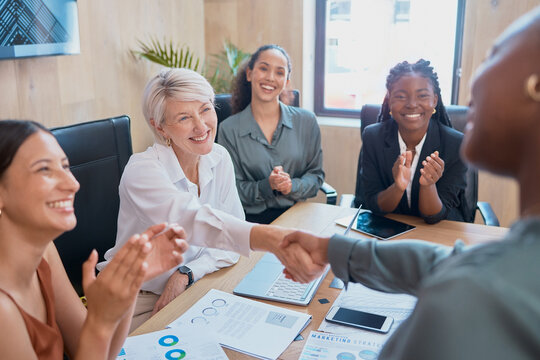Handshake between business women. Two diverse colleagues shaking hands during a meeting in the boardroom. Congratulating her on a job well done while coworkers clap in acknowledgement of achievement