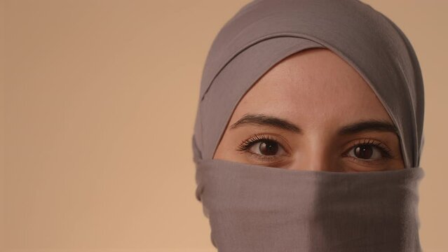 Close up studio portrait of brown eyed Muslim woman in niqab looking at camera standing on pastel background
