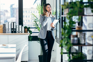 Beautiful elegant business woman talking with smartphone while holding some notes standing next to the window in a modern office