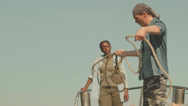 Multiethnic Couple Of Sailors Talking While Working Together On Boat In Sunlight. Man Throwing Anchor Rope In Water Standing On Deck