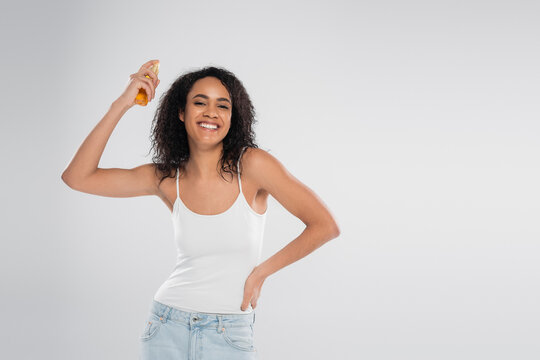 smiling african american woman applying hair oil while standing with hand on hip isolated on grey.