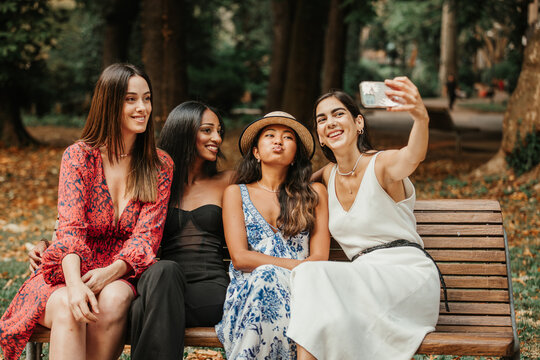 Four friends are sitting on a patch in a public park and taking a selfie together. ethnic friendship between women. - Powered by Adobe