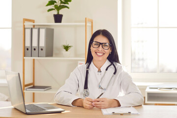 Portrait of cheerful young friendly female doctor in white medical uniform sitting and working on laptop computer. Happy woman physician in stethoscope looking at the camera and smiling.