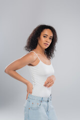 brunette african american woman in white tank top holding hand behind back while looking at camera isolated on grey.