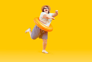 Fototapeta na wymiar Funny happy overweight woman having fun on summer holiday. Cheerful joyful fat girl in Tshirt, shorts, beach floater ring, sun hat and sunglasses dancing isolated on yellow background Vacation concept