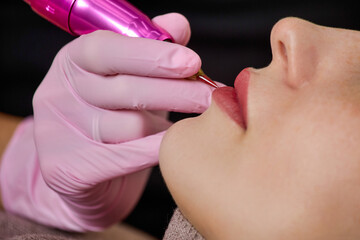 Macro photo of the process of professional permanent lip makeup with lipstick effect for a young woman. Close up, the hand of a master with a tattoo machine performing lip tattooing in a beauty salon - obrazy, fototapety, plakaty