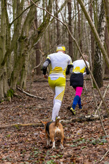 Young couple wearing sportswear and a dog running through a forest - obrazy, fototapety, plakaty