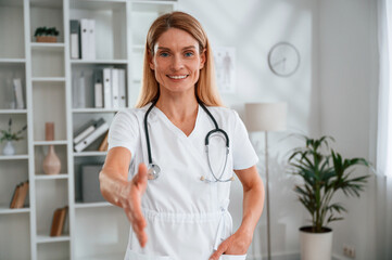 Giving hand to shake, greetings. Young female doctor in white coat is indoors