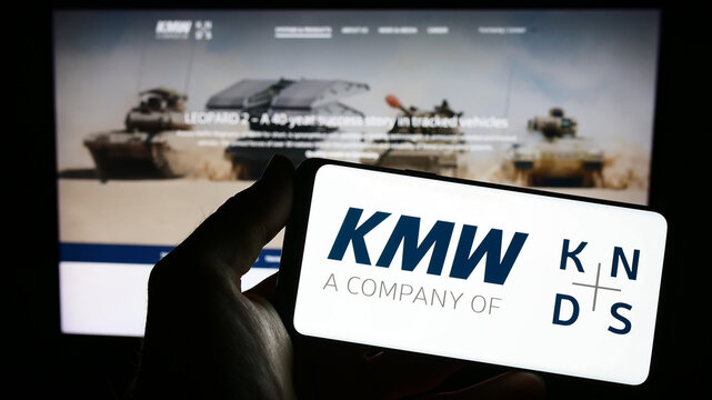 Stuttgart, Germany - 04-25-2023: Person holding smartphone with logo of Krauss-Maffei Wegmann GmbH Co. KG (KMW) on screen in front of website. Focus on phone display.