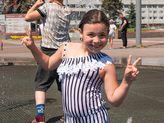 A little girl wet smiles holding up two fingers, happy bathing on a hot day in the city fountain....