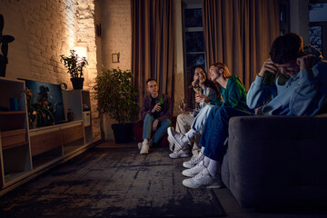 Obraz na płótnie Canvas Group of friends, young people sitting on couch at home in the evening and singing karaoke, having fun, talking and drinking beer. Concept of friendship, leisure activity, weekends, fun, emotions