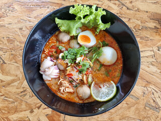 Creamy Tom Yum Noodles In a black bowl served with squid, prawns, meatballs, lettuce and boiled egg. Close-up shot focusing on selected point.