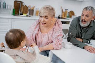 happy woman holding bowl near granddaughter eating breakfast in kitchen while bearded man opening mouth on blurred background.