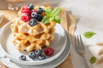Traditional Belgian waffles stacked with fresh raspberries and blueberries sprinkled with powdered sugar on the kitchen counter