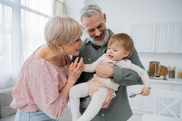 pleased middle aged couple playing with little granddaughter holding spoon in kitchen.
