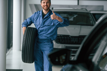 Walking forward and holding the tire. Man in blue uniform is working in the car service