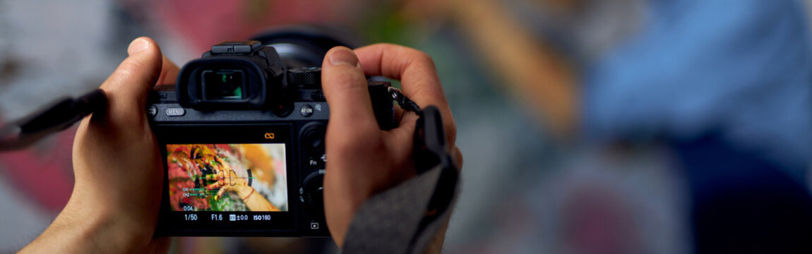 Close up of male hands holding professional digital camera on a blurred background. Female artist creating painting in the background