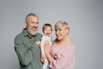 Fototapeta na wymiar happy mature couple looking at camera near laughing granddaughter isolated on grey.