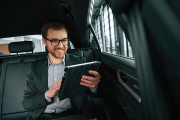 Smiling, using tablet. Man in formal business clothes is sitting in the modern automobile