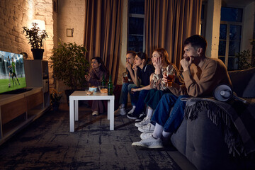 Obraz na płótnie Canvas Group of young people, friends sitting on couch at home in the evening and watching live football match translation on tv. Concept of friendship, leisure activity, weekends, fun, emotions