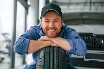 Leaning on the tire and smiling. Man in blue uniform is working in the car service