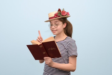 A girl in retro style clothes enthusiastically reads a book a straw hat with flowers, glasses and a...
