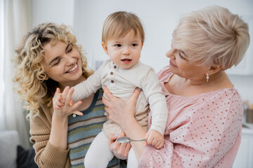 joyful women looking at toddler girl holding spoon in kitchen.