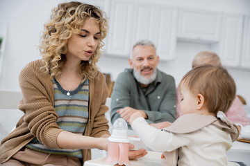 Obraz premium woman feeding little daughter with breakfast near mature parents smiling on blurred background in kitchen.