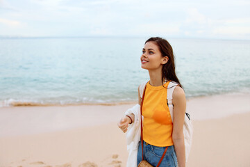 Woman smile with teeth after swimming in the ocean with a backpack in a wet yellow tank top and denim shorts walks along the beach, summer vacation on an island by the ocean in Bali sunset