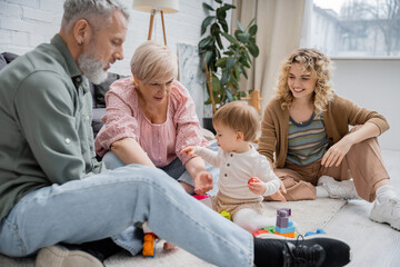 joyful woman looking at middle aged parents playing building blocks game with child on floor in living room.