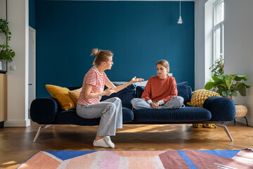 Upset teen girl sitting on sofa listening angry mother scolding her for bad school grades. Mom...