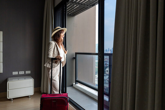 Asian woman moving the luxury hotel with luggage on her vacation while looking at the view from window with high rise building in the urban downtown area for business trip and condominium living usage